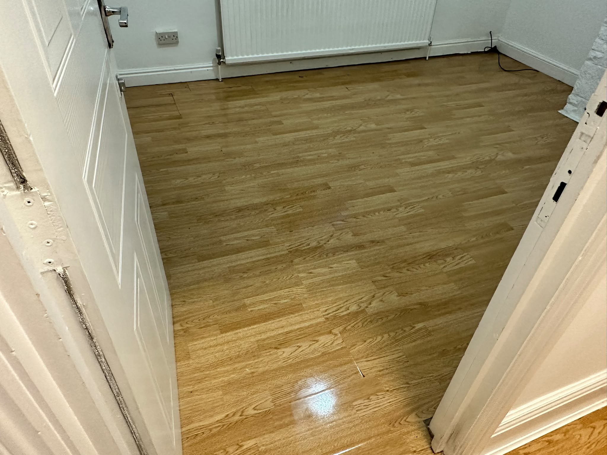 Empty room with a polished light wood laminate floor, white door and radiator, seen from the doorway